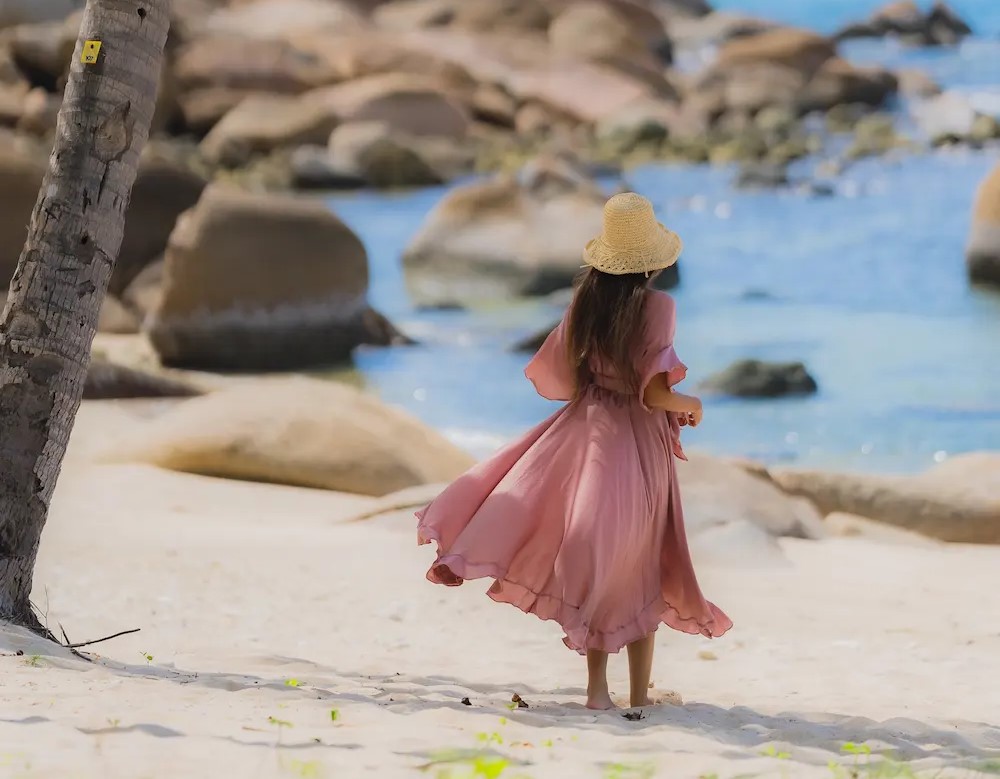 Woman in pink dress and straw hat walking on a sandy beach with rocks and blue water