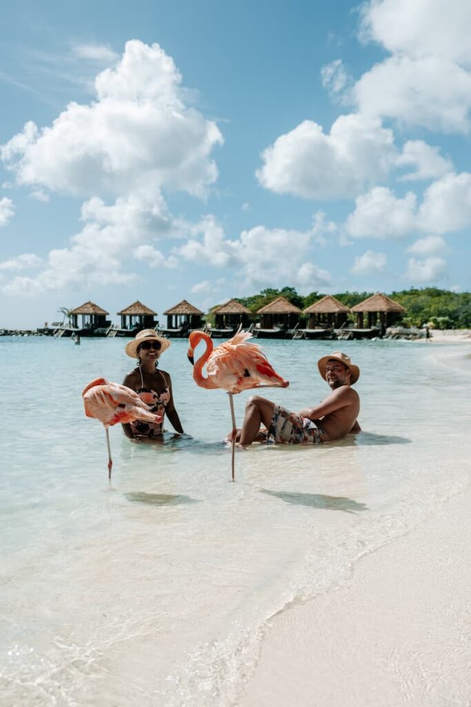 Two people sitting in shallow turquoise water with flamingos, overwater bungalows and blue sky with clouds in the background